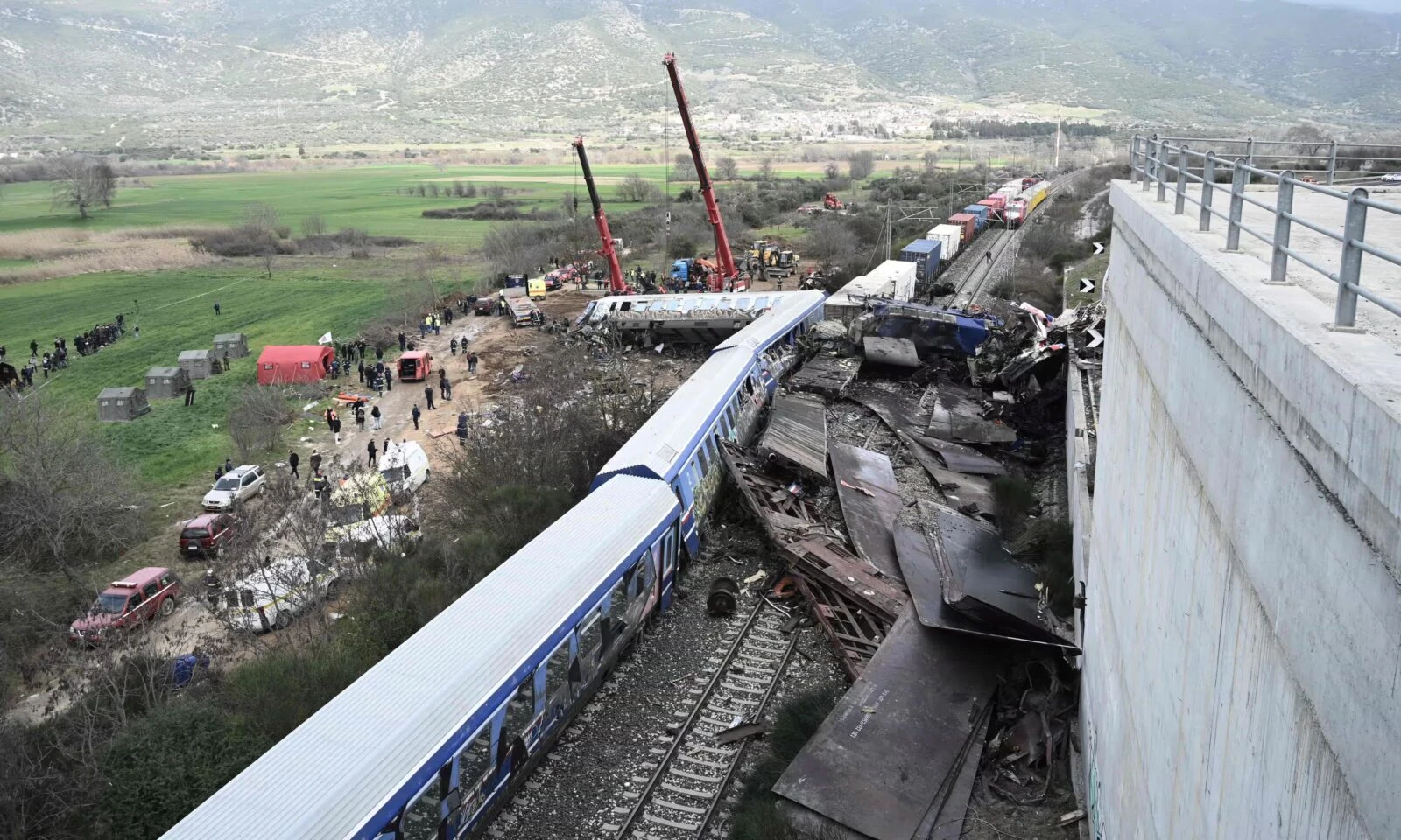 A close-up of the wreckage from the train collision in Tempi, Greece. Crushed train parts and debris are piled up near a concrete barrier, while rescue workers in safety vests inspect the scene.