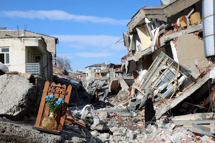 A small decorative "Home" sign with blue flowers sits on top of debris, surrounded by crumbled buildings after February 6 earthquake