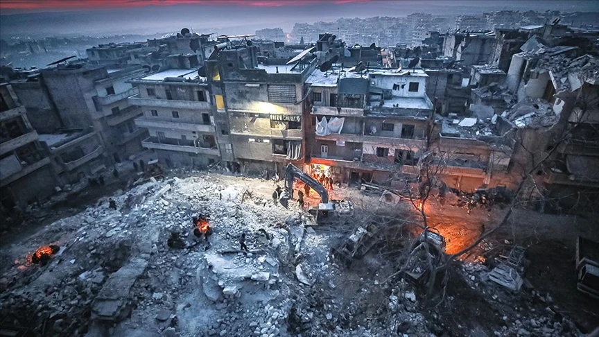 An aerial view of destroyed buildings at night, with searchlights and rescue workers digging through the rubble in the dark