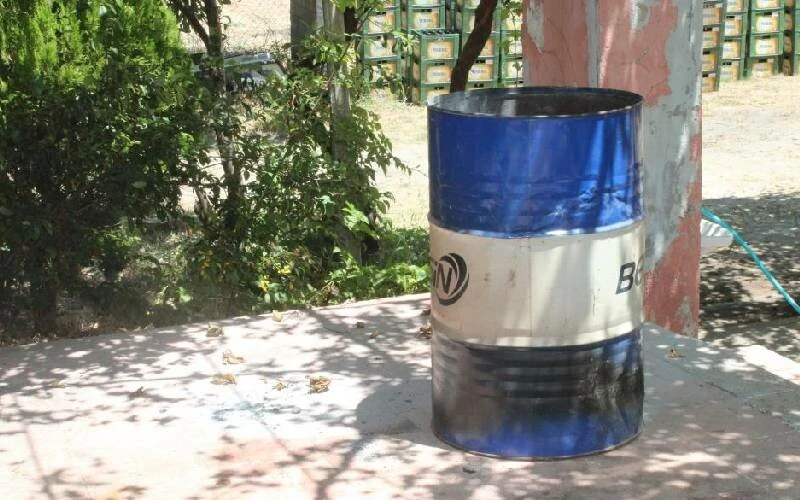 A blue and white metal barrel, partially burned, standing outdoors with trees and plants in the background