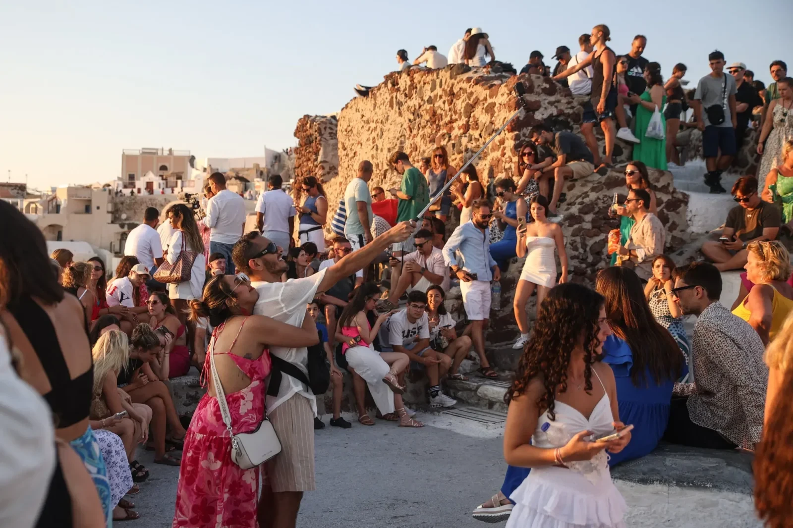 A crowd of tourists enjoying the sunset on Santorini, a popular yet overtouristed Greek island, with its iconic white-washed buildings and blue-domed churches in the background.