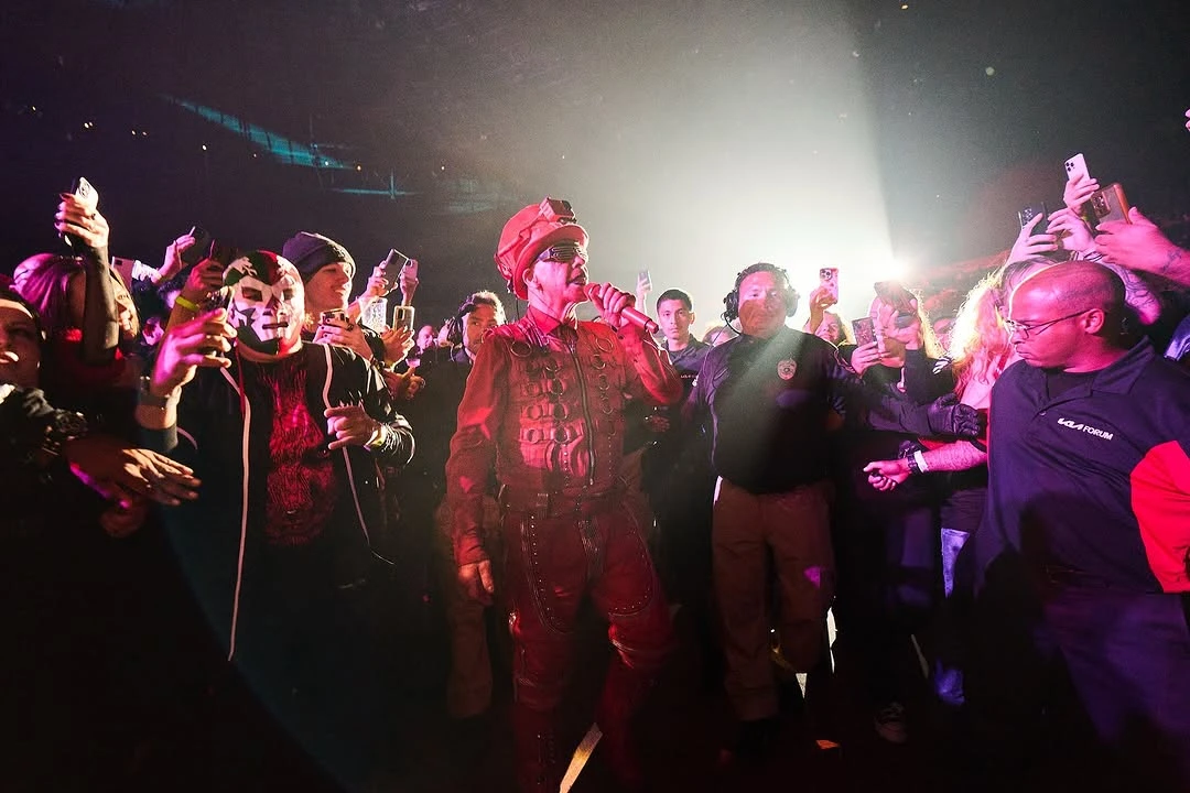 Till Lindemann, dressed in a red military-style outfit and sunglasses, walks through a crowd during a live performance, holding a microphone. Fans surround him, taking photos and videos with their phones, while security personnel ensure his path remains clear in a dimly lit venue with dramatic lighting.