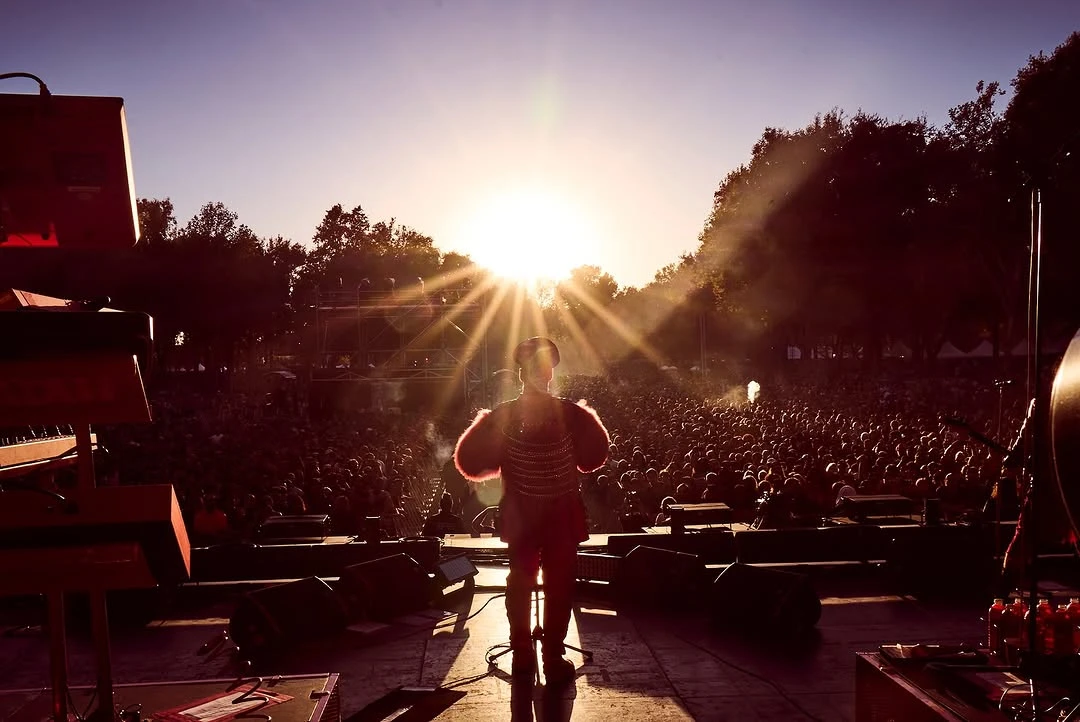 Till Lindemann stands on stage at an outdoor concert, facing a massive crowd as the sun sets behind him. Wearing an elaborate red outfit with fur-trimmed sleeves, he is illuminated by golden sunlight streaming through the trees, creating a striking silhouette against the audience and stage equipment.