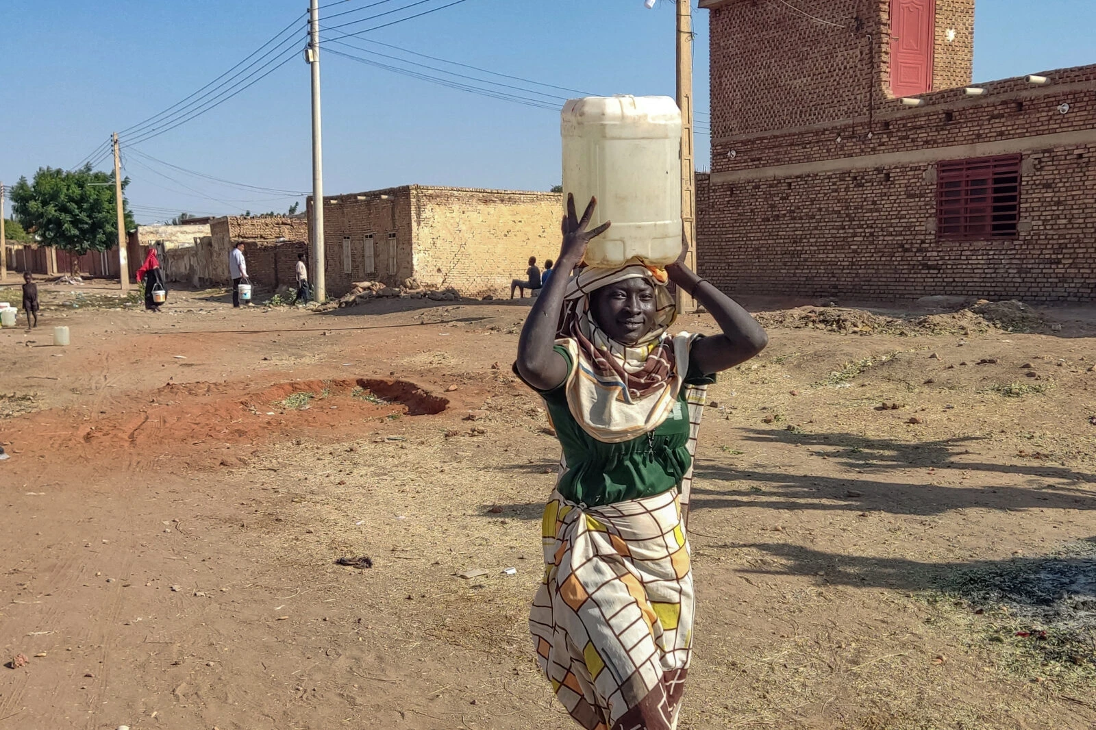 A woman smiles while carrying a large white water container on her head in a dusty neighborhood in Sudan. She wears a green top and patterned wrap skirt, standing in an unpaved street surrounded by brick buildings. In the background, other people can be seen carrying water containers, highlighting the daily water collection routine in this arid community. Power lines stretch across the clear blue sky above the settlement's simple structures.