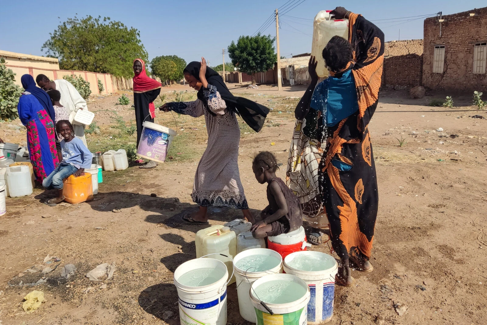 People gather water in a dusty street in Sudan. Women and children stand around multiple plastic containers and buckets filled with water. One woman carries a large water container on her head, while others tend to various containers. They're dressed in colorful traditional garments against a backdrop of simple brick buildings and sparse vegetation, illustrating the daily struggle for water access in what appears to be a resource-limited environment.