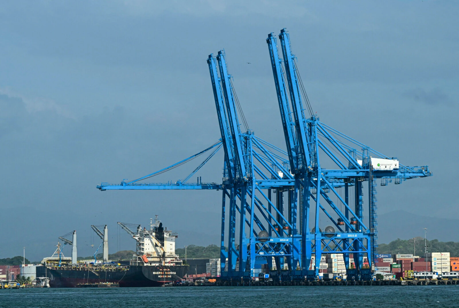 The image shows a port with large blue gantry cranes towering over cargo containers and docked ships