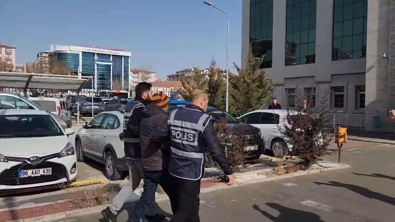 A man in handcuffs wearing a beanie is escorted by two police officers in vests, while a cameraman films the scene in a busy urban area with parked cars and buildings.