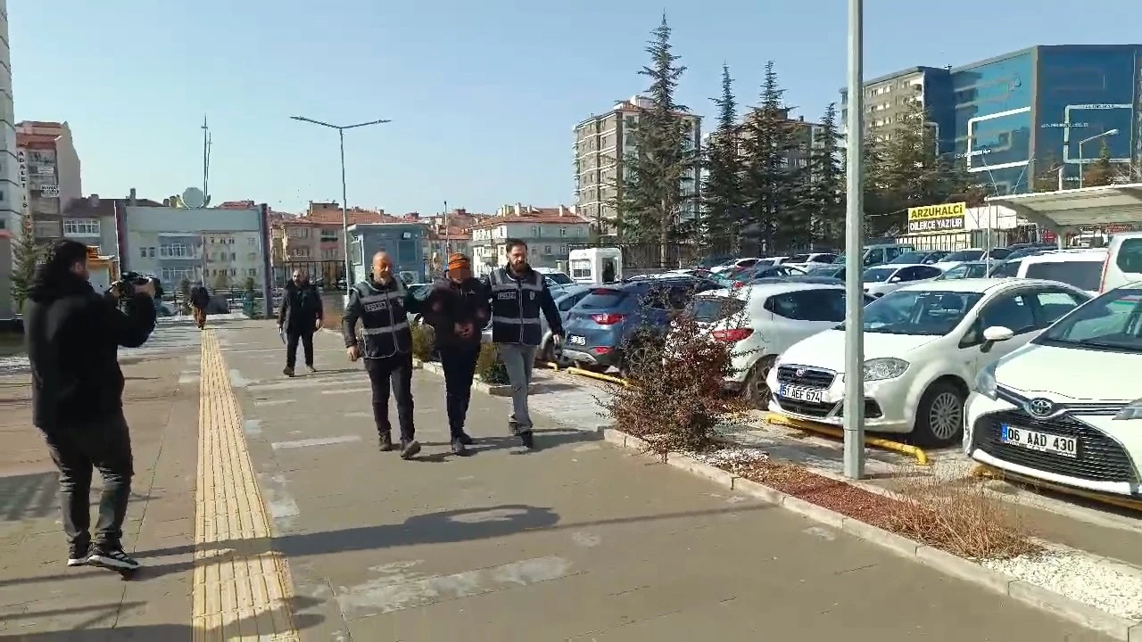 A man in handcuffs wearing a beanie is escorted by two police officers in vests, while a cameraman films the scene in a busy urban area with parked cars and buildings.