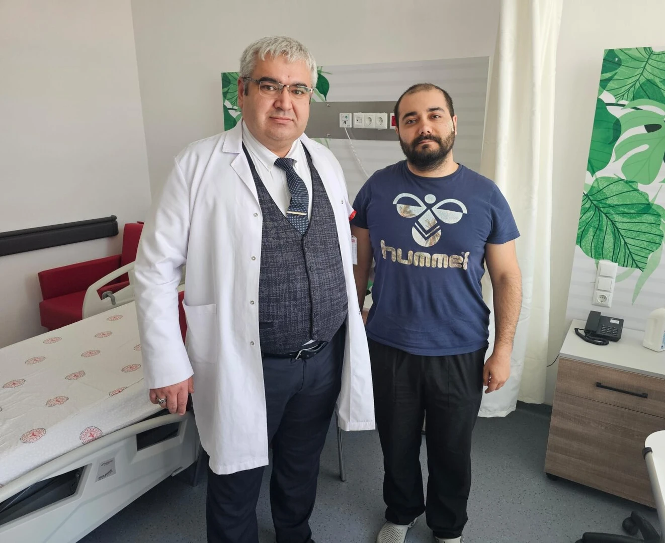 A doctor in a white coat stands next to a man in a blue t-shirt inside a hospital room, with a bed and green wall decor visible in the background.