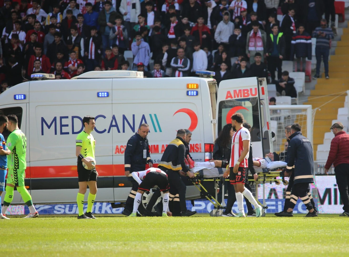 An ambulance is parked on the sideline of a football stadium as medical personnel carry a footballer