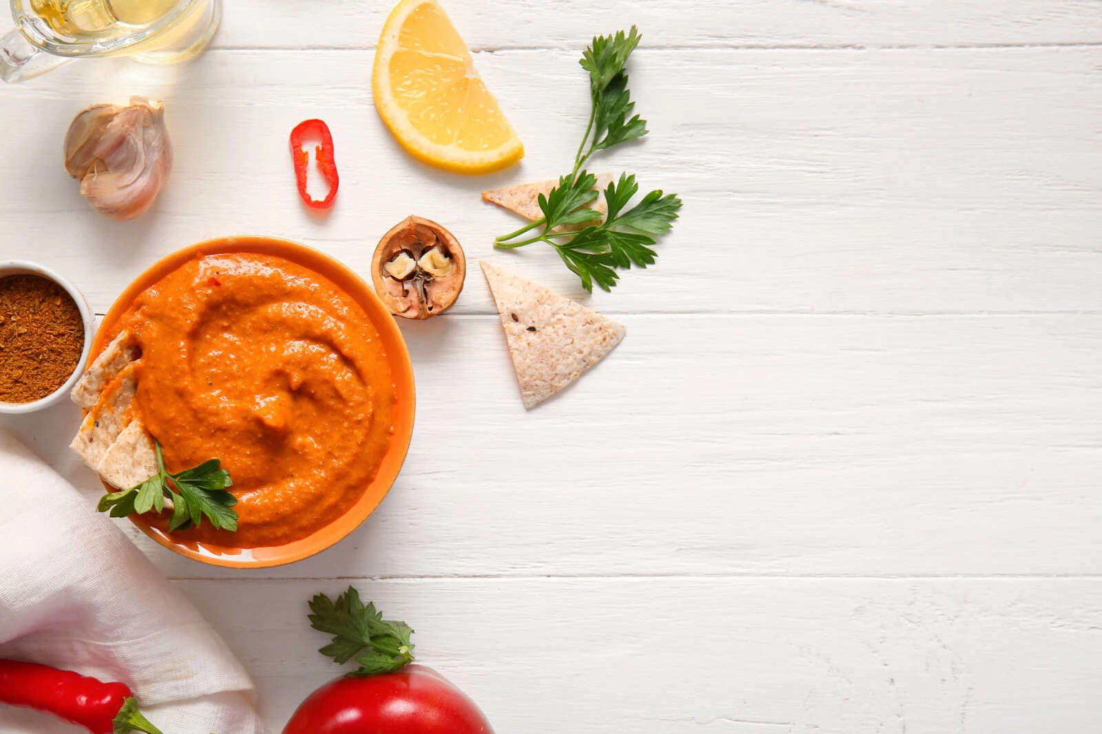 A vibrant red pepper and walnut dip garnished with fresh parsley, surrounded by lemon wedges, walnuts, and pita chips.