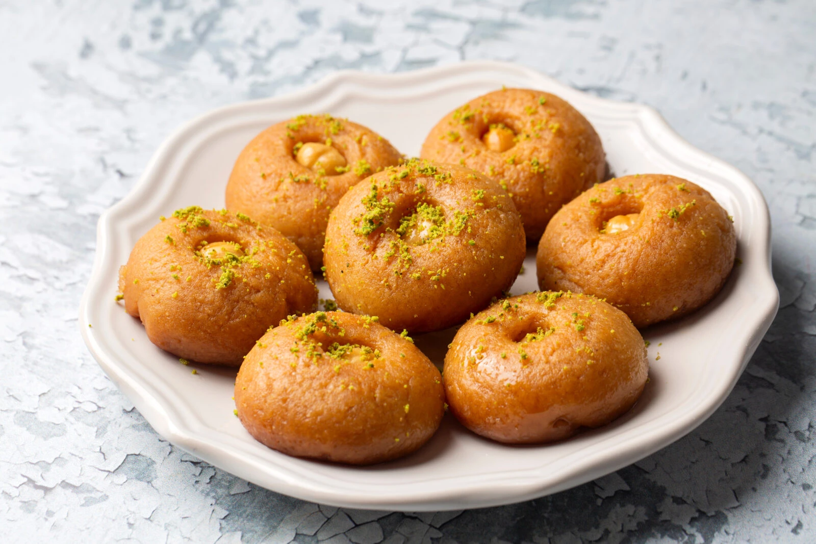 A plate of Turkish Sekerpare, golden-brown semolina cookies soaked in syrup, topped with crushed pistachios, arranged on a decorative white dish.