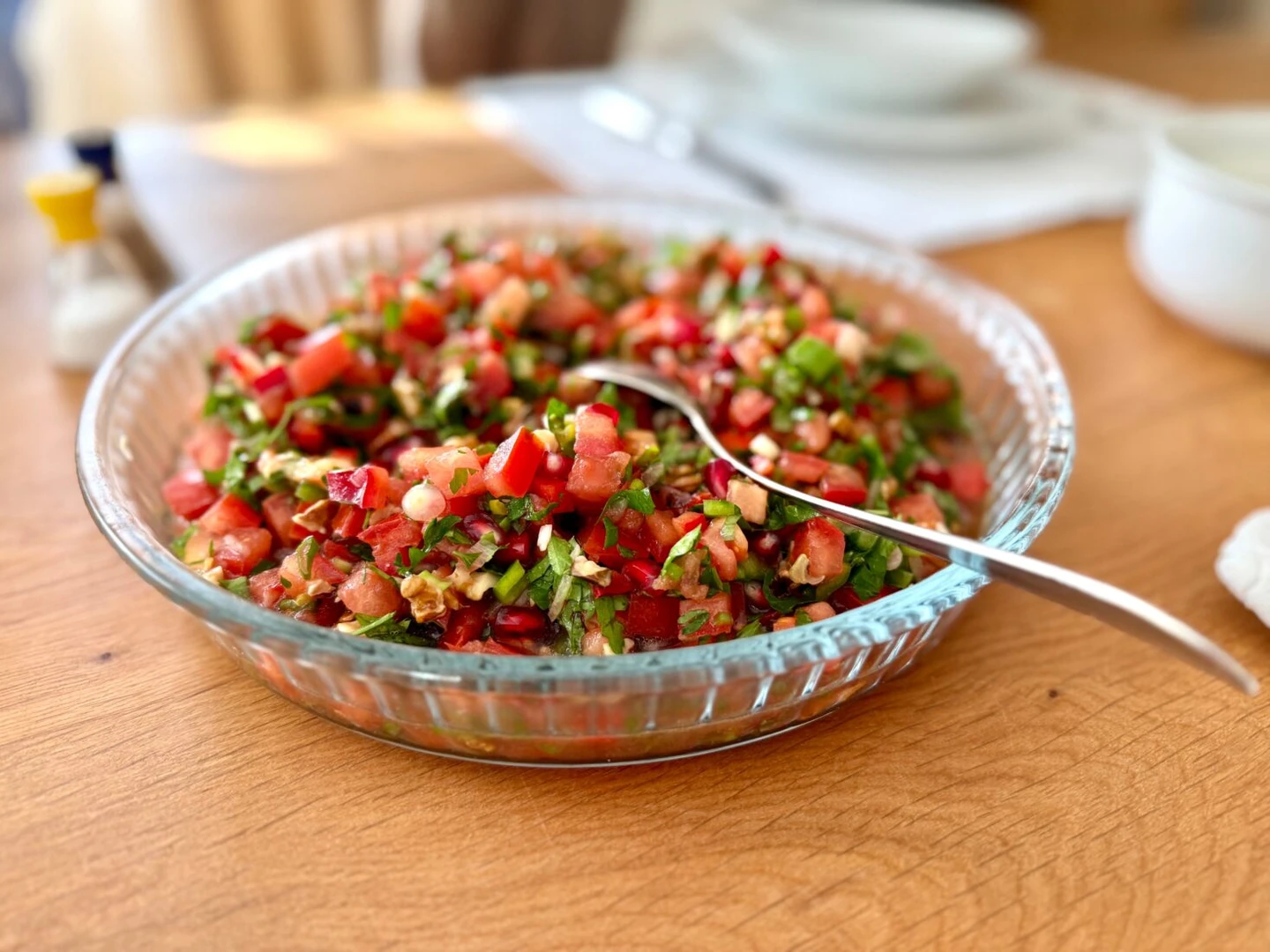 Break fast in ramadan - A glass bowl filled with Gavurdağı salad, a Turkish chopped salad made with tomatoes, cucumbers, onions, and walnuts, with a spoon resting inside. The background features a wooden table with salt and pepper shakers.