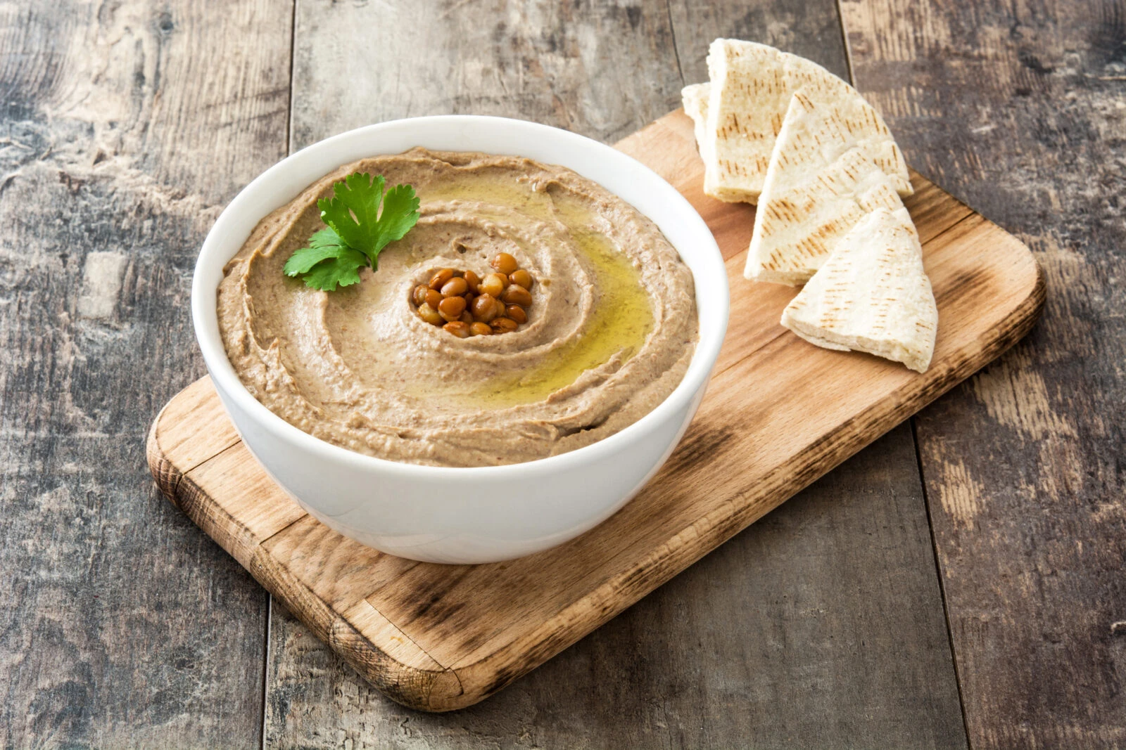 A bowl of creamy red lentil dip garnished with olive oil and whole lentils, served with pita bread on a wooden board.