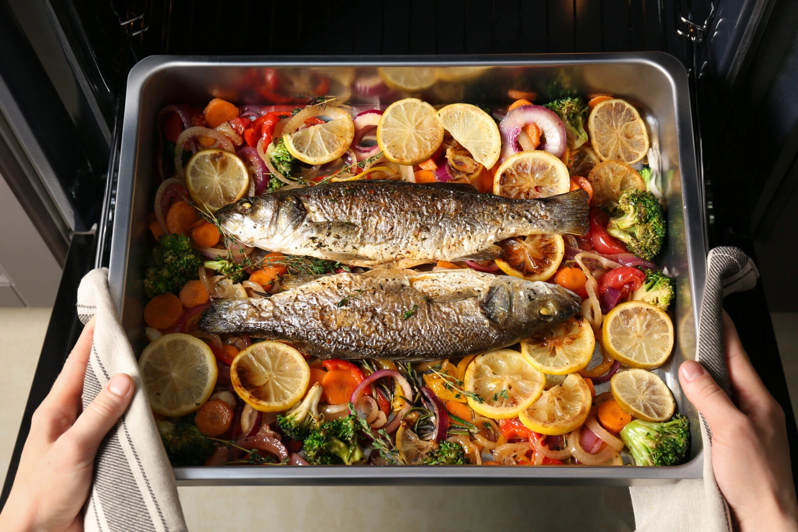 Two whole fish in a baking tray, surrounded by onions, tomatoes, broccoli, and lemon slices, just out of the oven.