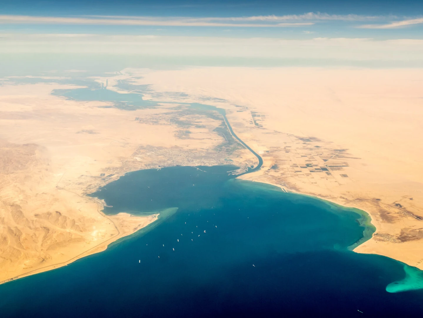 An aerial view of the Suez Canal, showing its connection between the Mediterranean Sea and the Red Sea, surrounded by desert landscapes.