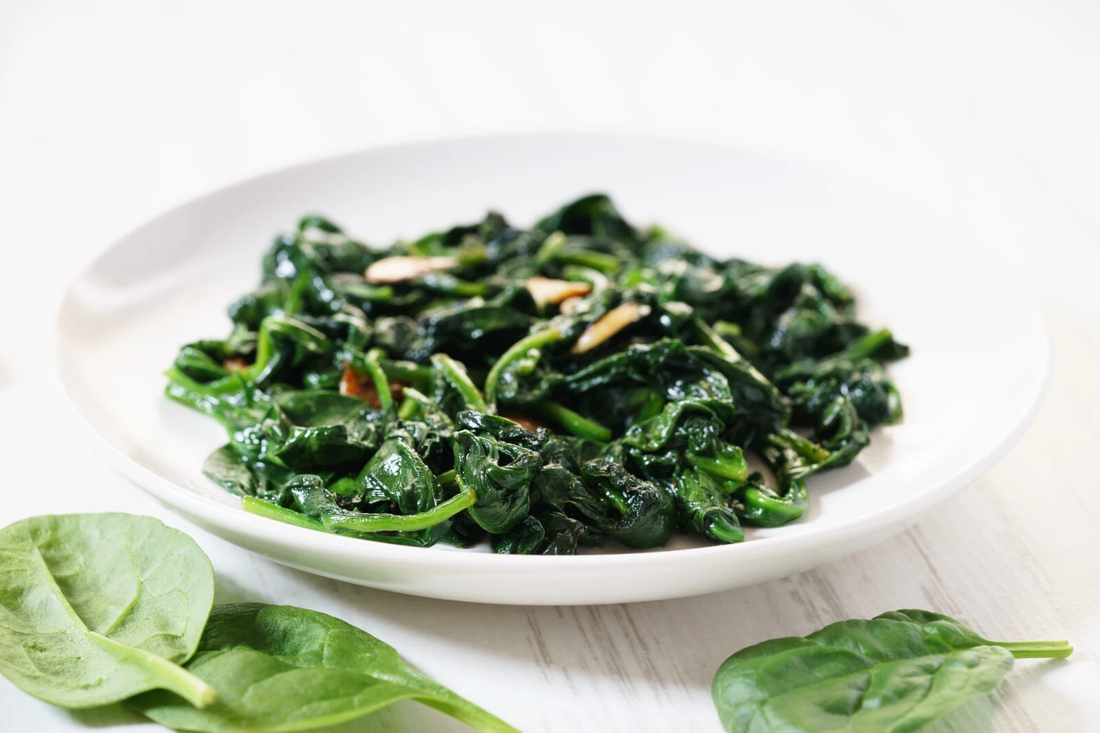 A plate of sautéed spinach with garlic, served on a white dish with fresh spinach leaves in the background.