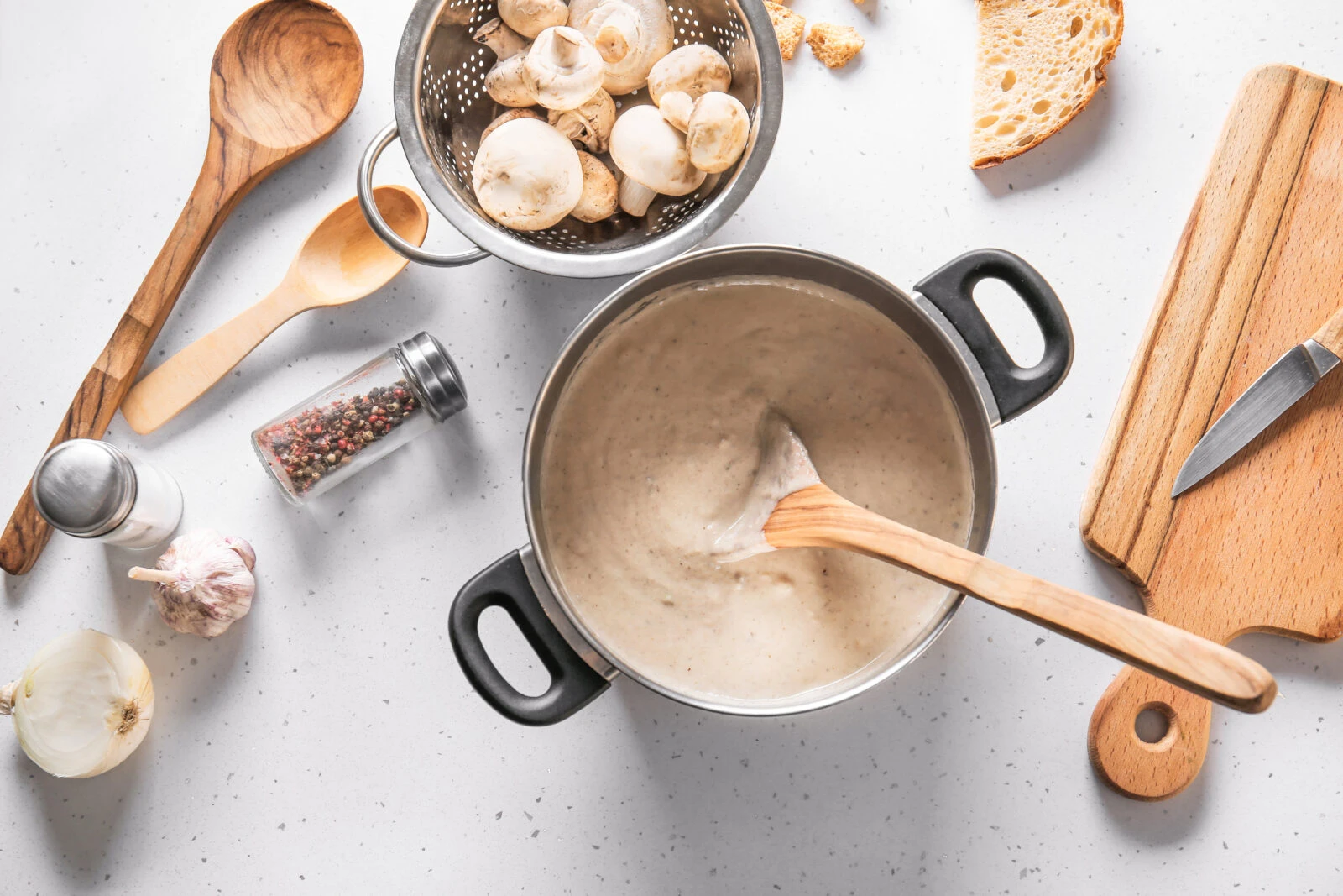     A pot of creamy mushroom soup with a wooden spoon, fresh mushrooms, and seasoning ingredients on a kitchen counter.