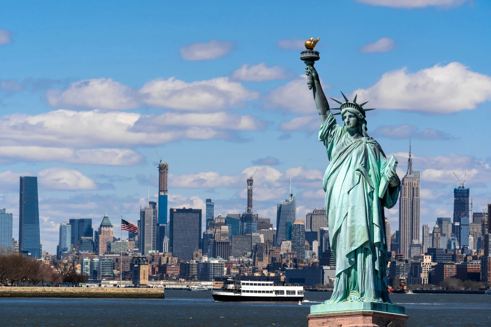 The Statue of Liberty with the Manhattan skyline and One World Trade Center in the background on a clear day.