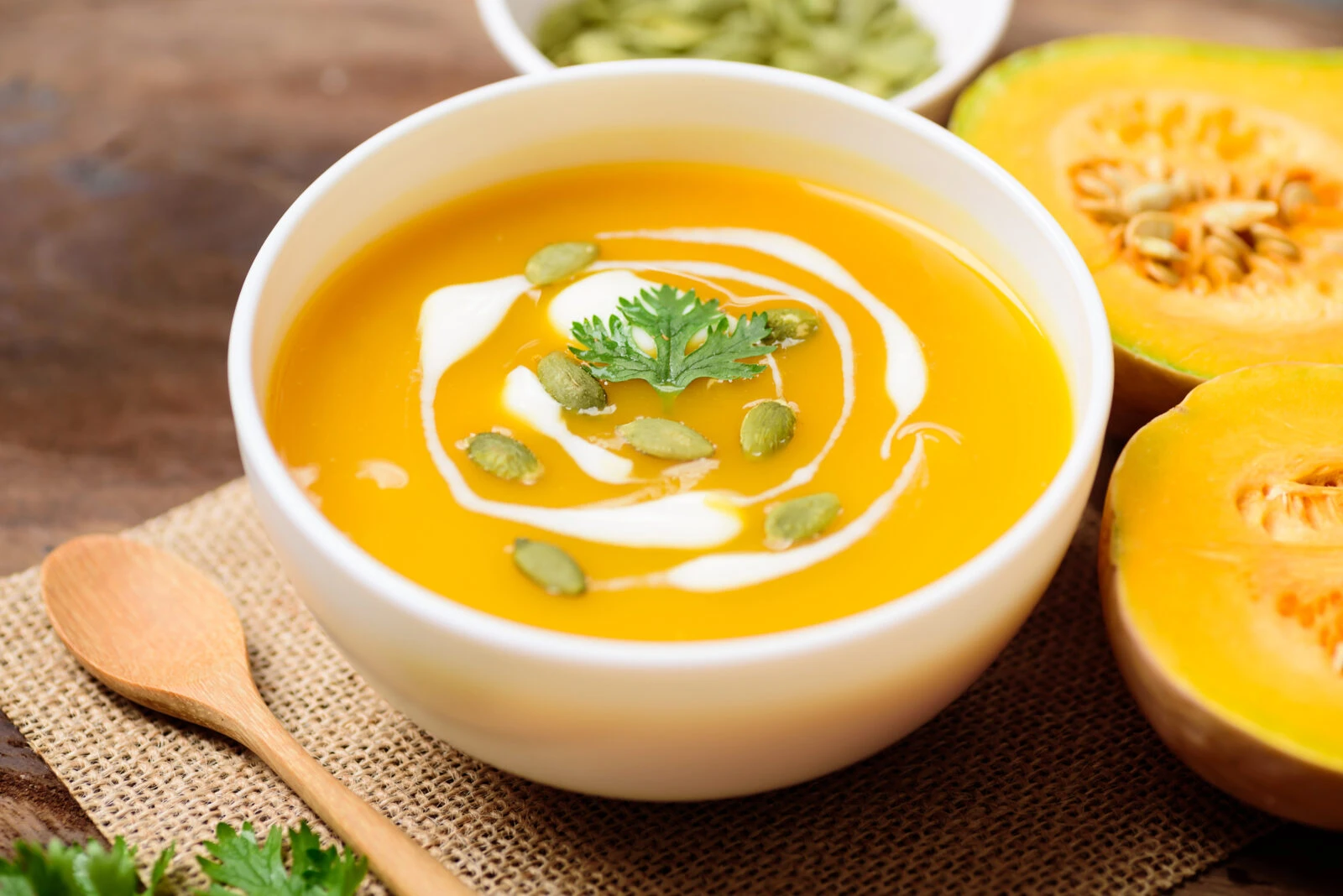 A steaming bowl of creamy pumpkin soup with a swirl of cream, garnished with pumpkin seeds and fresh herbs.