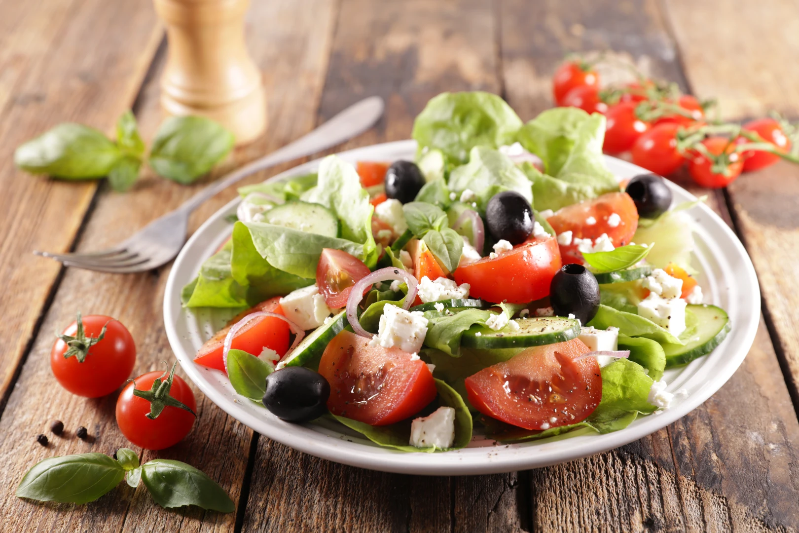 A plate of Mediterranean salad with lettuce, cherry tomatoes, cucumbers, black olives, and feta cheese on a rustic wooden table, garnished with fresh basil and a drizzle of olive oil.