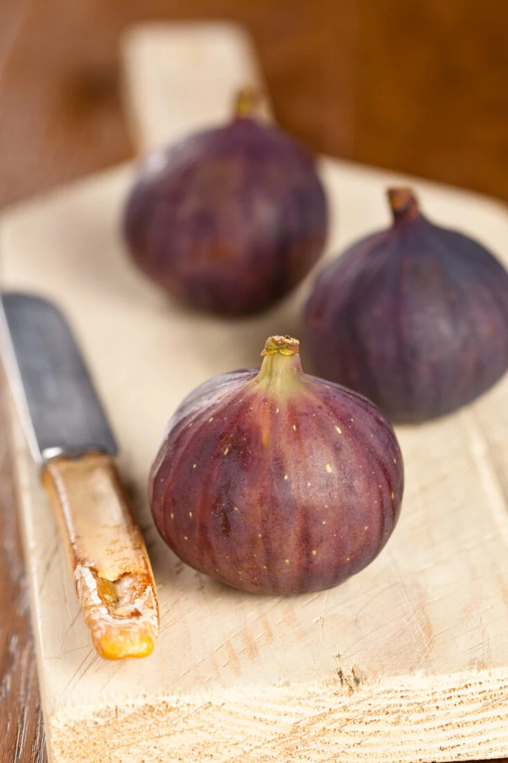 A close-up of black figs on a cutting board, one cut open to reveal its red interior, with a knife beside them.