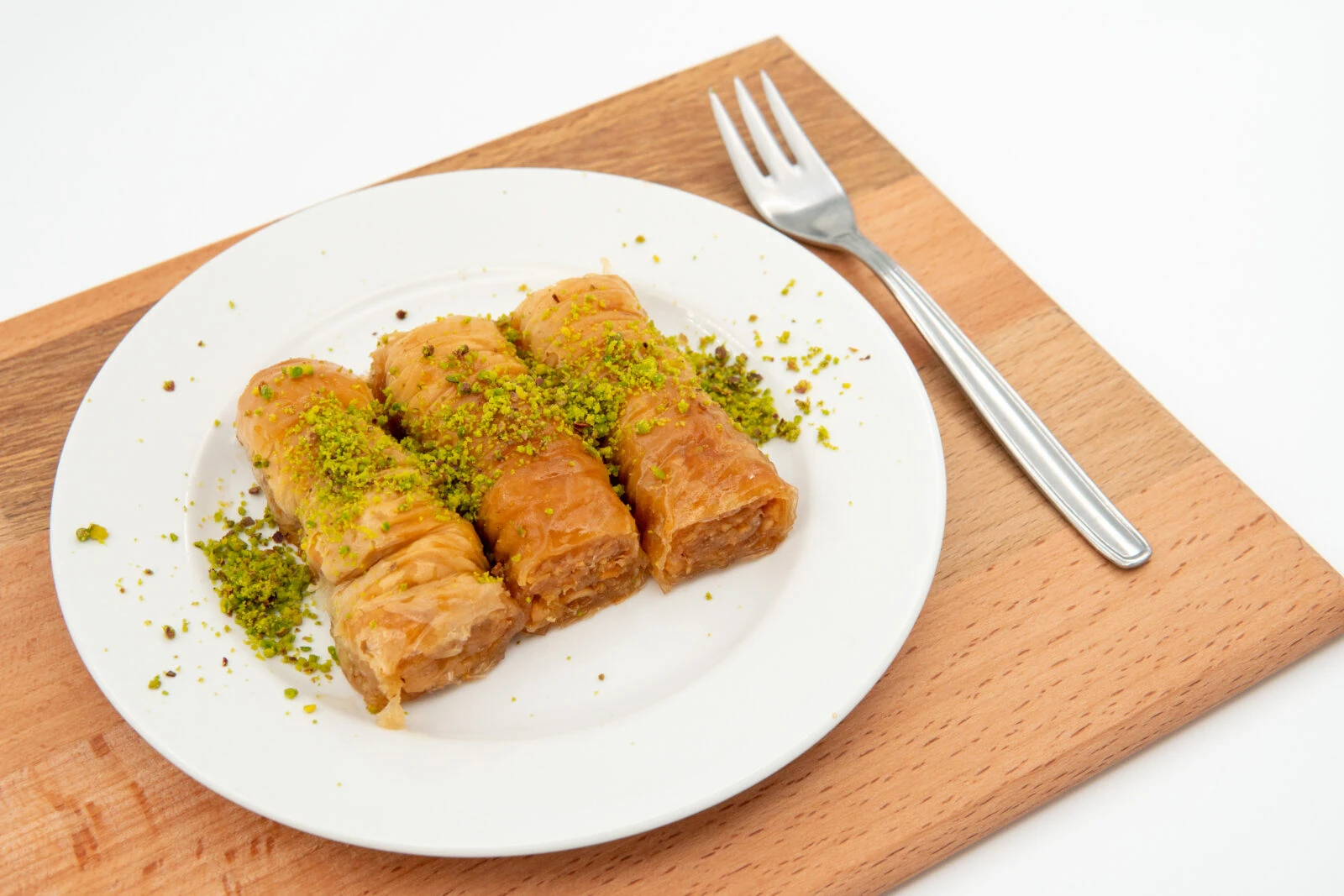 A plate of Turkish walnut-stuffed baklava rolls topped with crushed pistachios, placed on a wooden board with a silver fork.