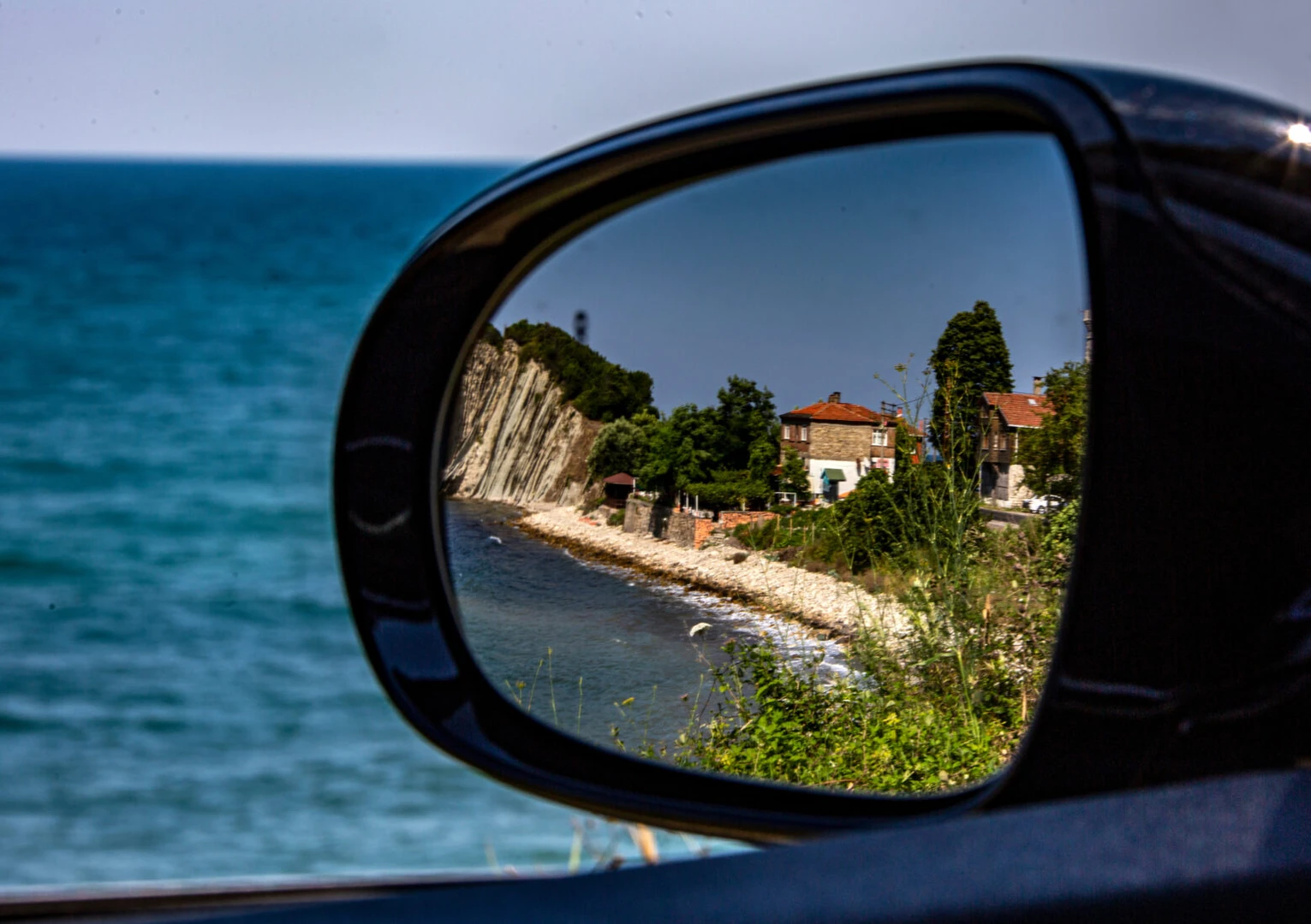 A coastal village near Kiyikoy reflected in a car’s side mirror, with the Black Sea and rocky cliffs in the background.