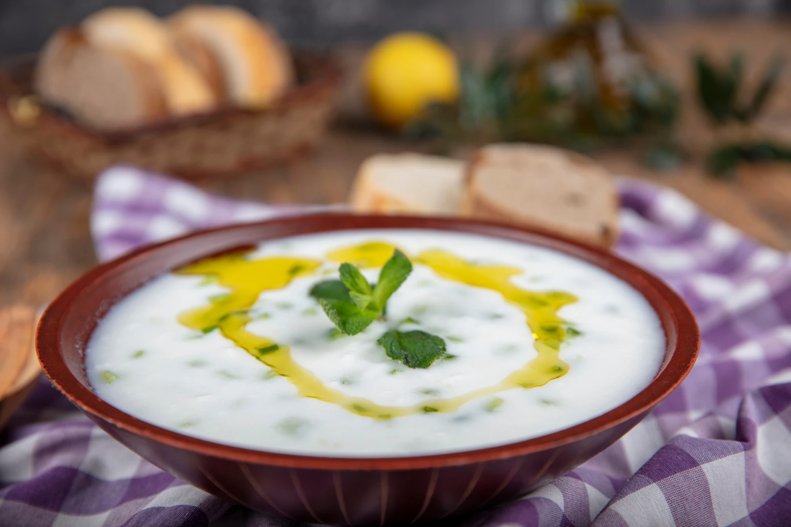 A bowl of Turkish cacik, a cold yogurt and cucumber dip, garnished with fresh mint and olive oil, served with bread in the background on a rustic table.