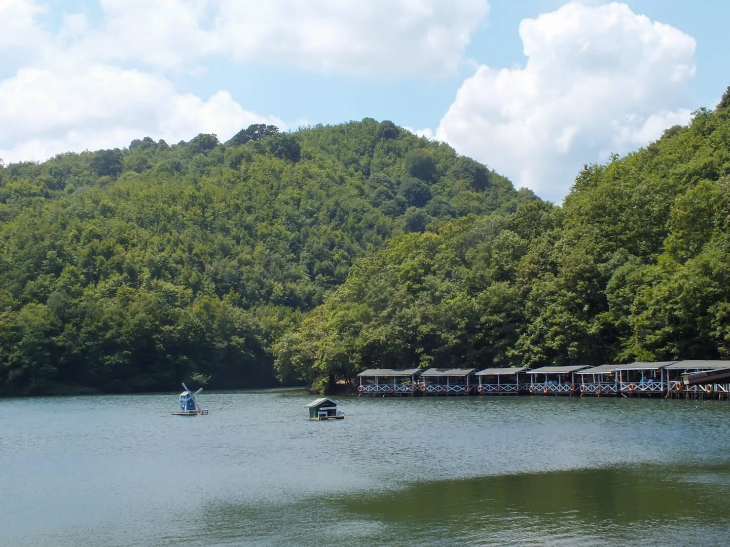 A view of Sakligol in Sile near istanbul, with a calm lake, floating windmill structures, and lakeside restaurants nestled in green forests.