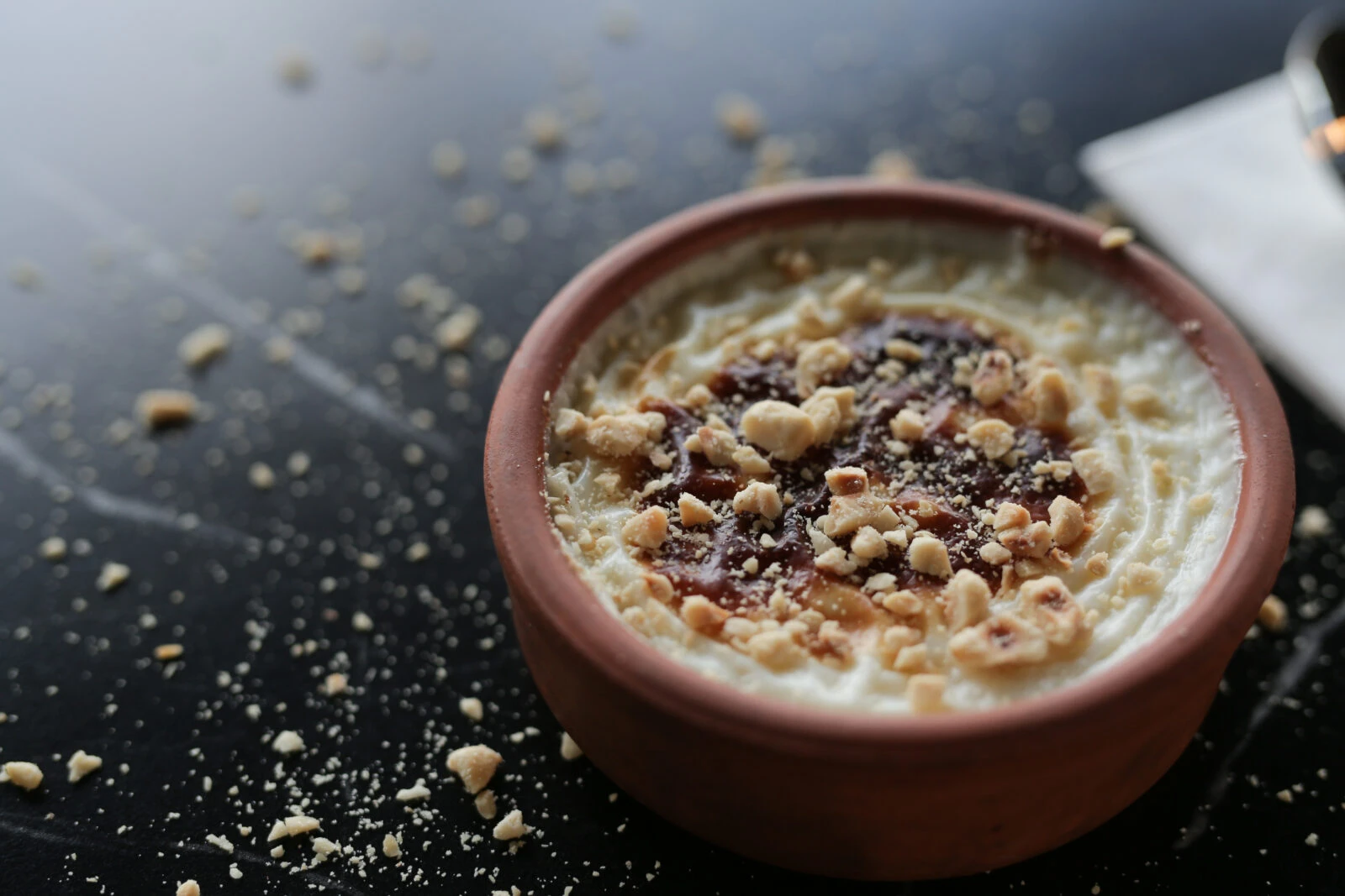 A bowl of traditional Turkish rice pudding with a caramelized top, garnished with crushed hazelnuts, placed on a black surface with scattered nuts.