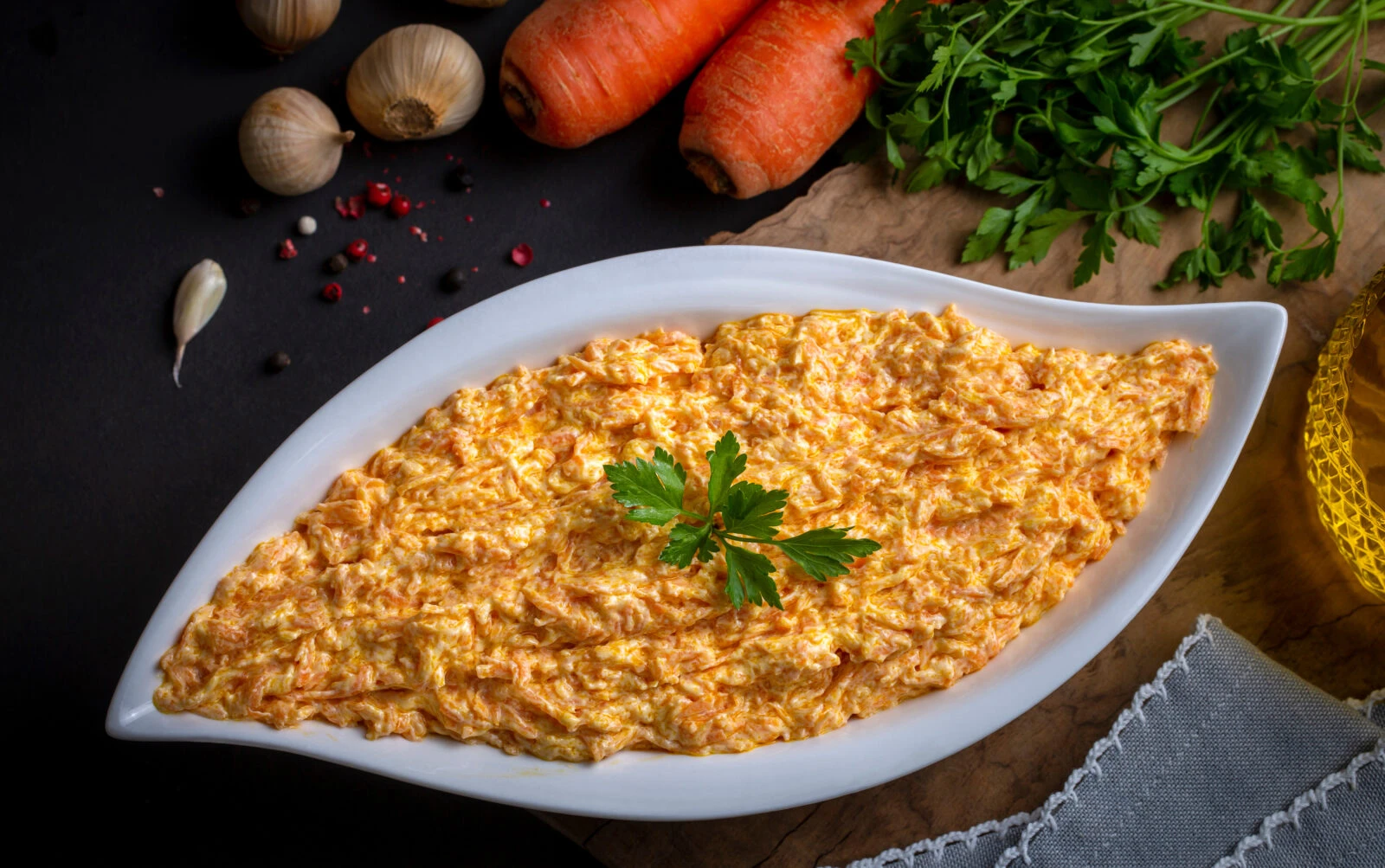 A white, curved dish filled with yogurt carrot tarator, a Turkish side dish made with grated carrots and yogurt, garnished with parsley. Whole carrots, garlic, and spices surround the dish on a rustic dark background.