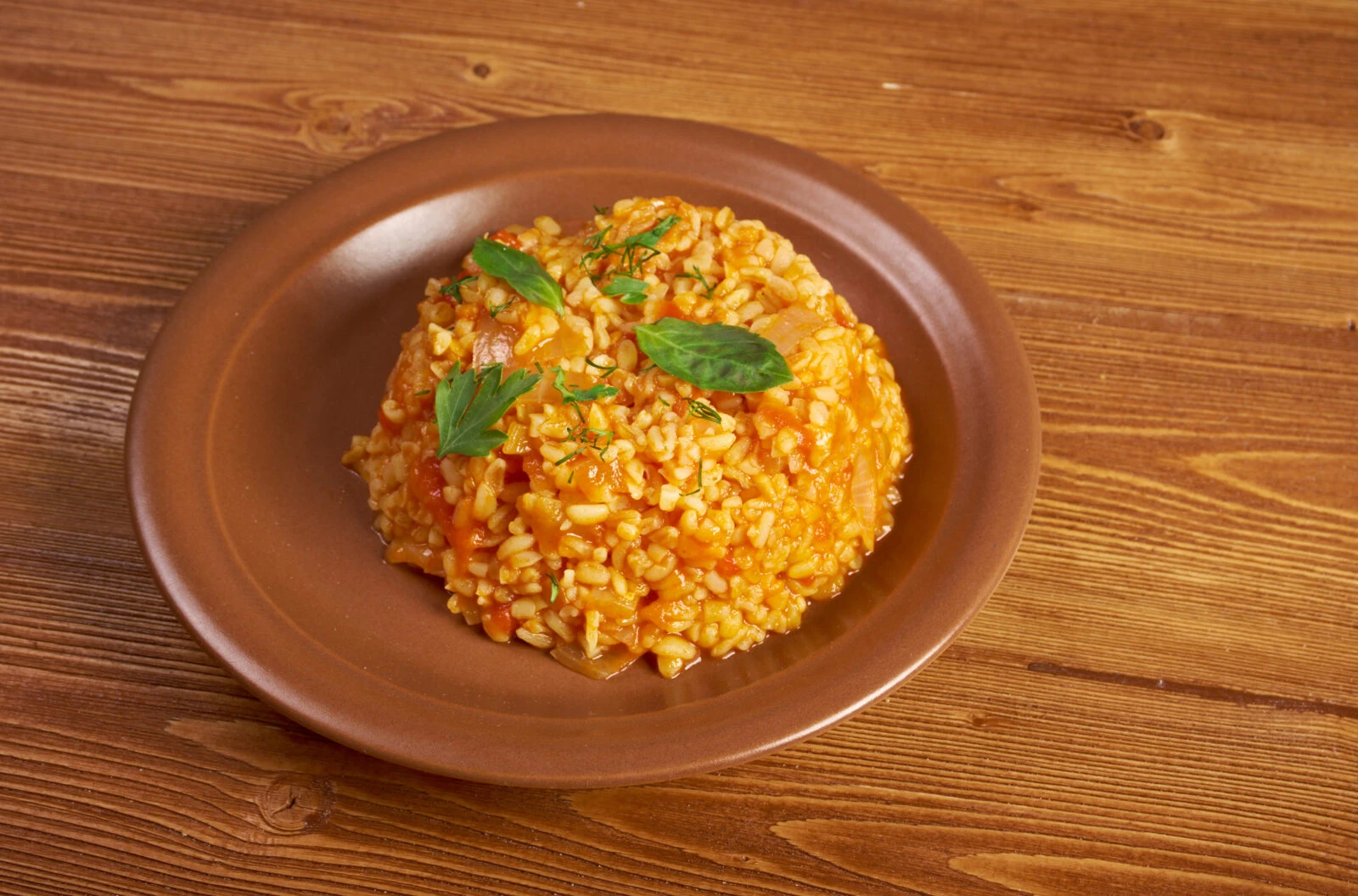 A plate of Turkish bulgur pilaf with roasted vegetables, garnished with fresh herbs, served on a rustic brown plate.