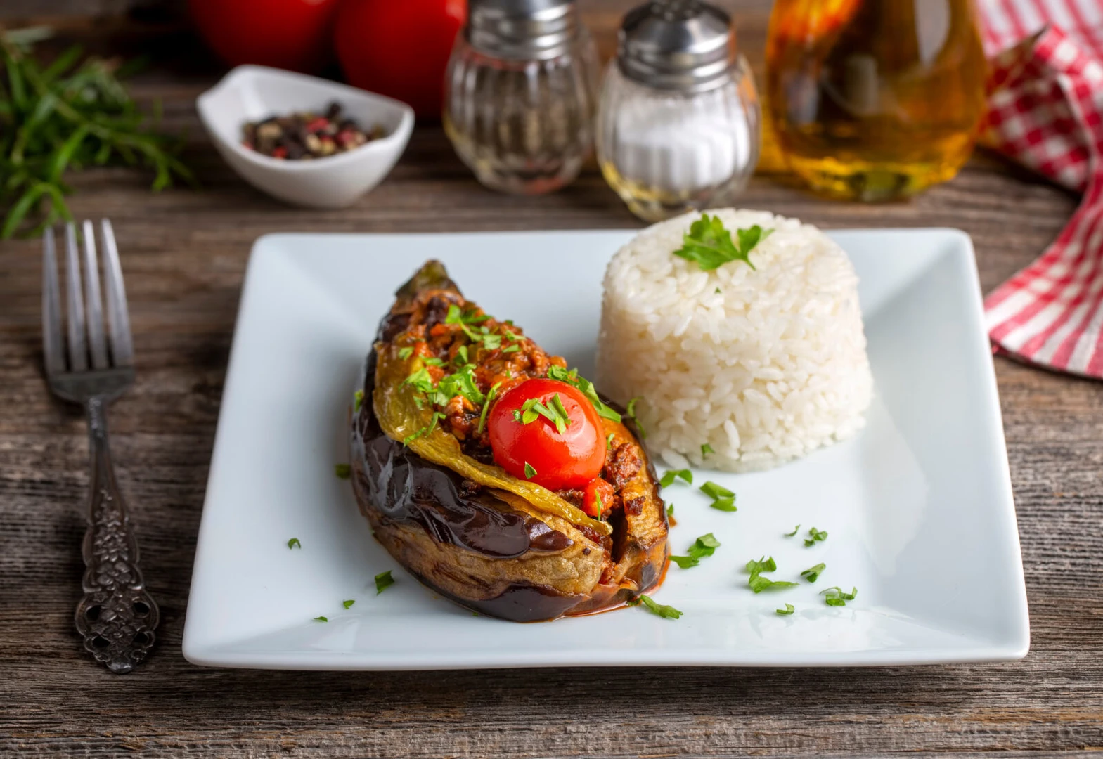 A plate of Turkish Karniyarik, featuring an oven-baked eggplant stuffed with seasoned ground beef, topped with a roasted tomato and green pepper, served with rice on a wooden table.