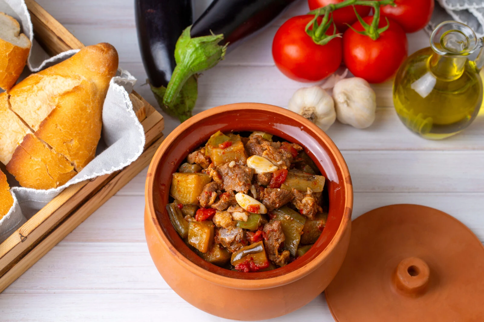 A terracotta dish filled with Turkish beef and vegetable casserole, surrounded by fresh tomatoes, garlic, eggplants, and bread.