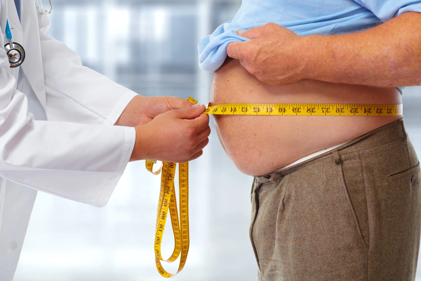 A doctor in a white coat using a yellow measuring tape to measure an overweight man's waist, highlighting concerns about obesity and health monitoring.