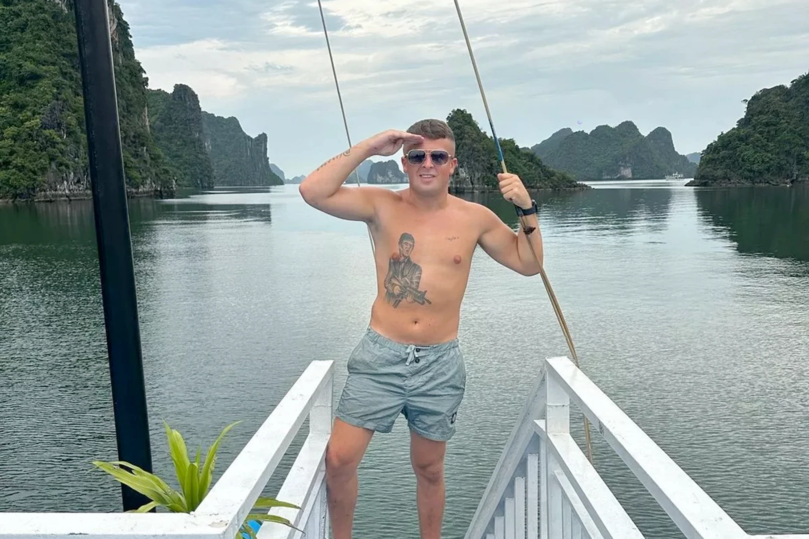 British fugitive Ewan Corbett stands on a boat deck, saluting with one hand while holding onto a railing with the other. The background features a calm bay with limestone islands.