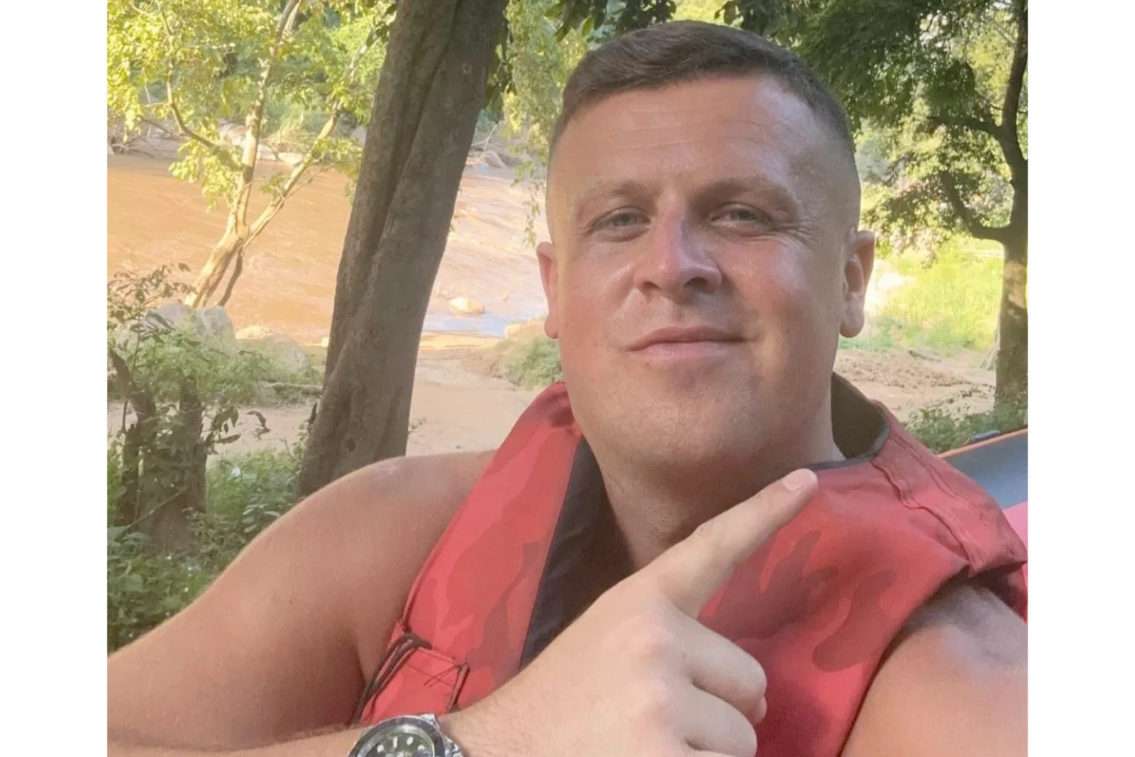 Ewan Corbett wears a red life jacket and points his index finger upward while taking a selfie outdoors, surrounded by trees and a muddy river.