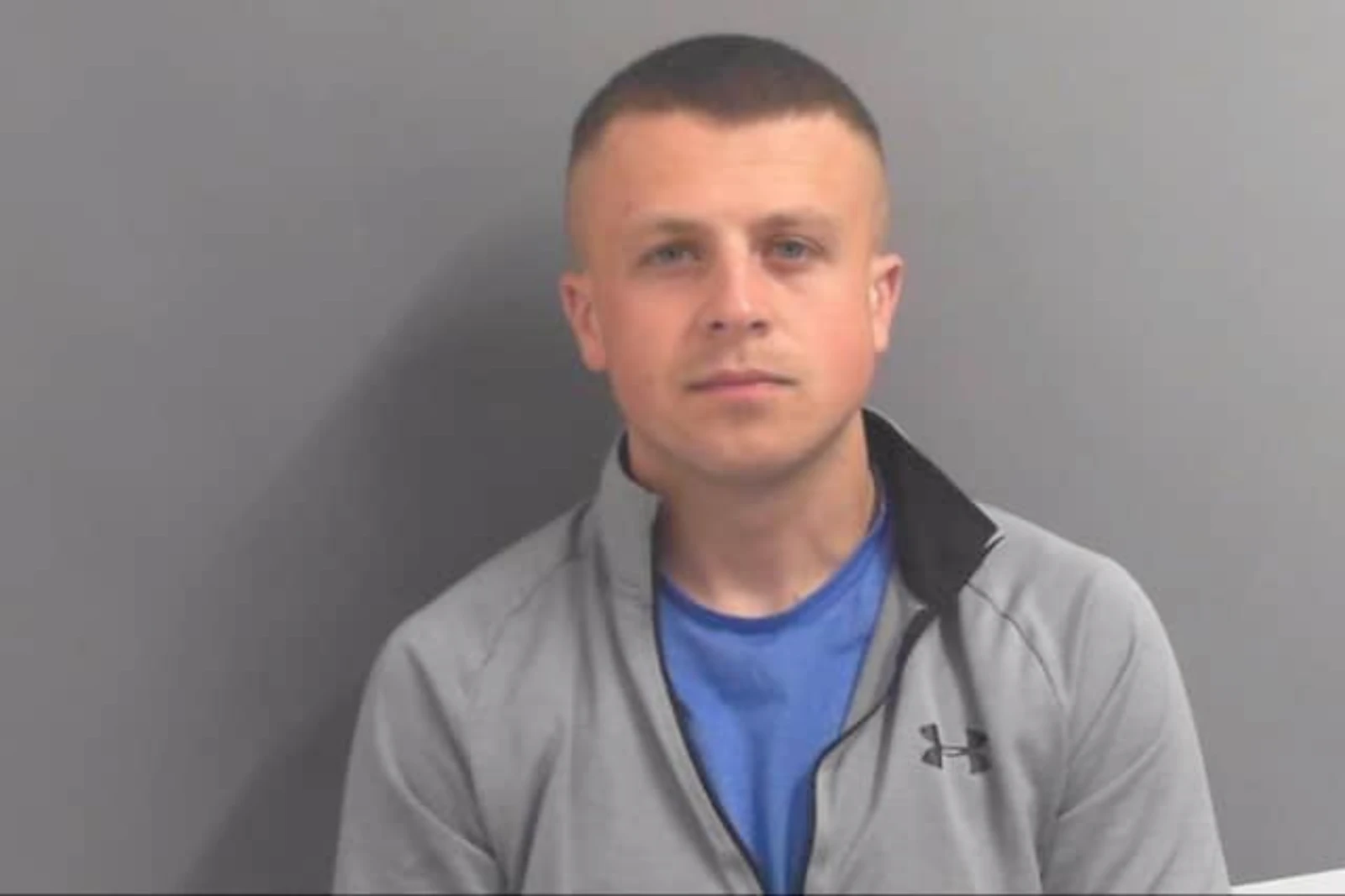 Official mugshot of British fugitive Ewan Corbett wearing a grey Under Armour zip-up jacket and a blue T-shirt, taken against a plain grey background.