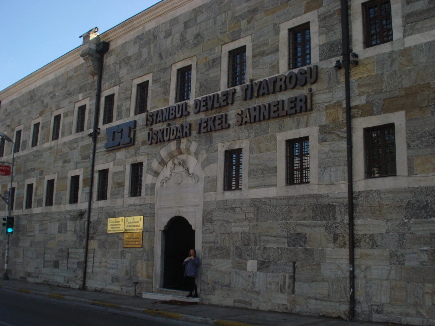 The stone facade of the Istanbul State Theater's Üsküdar Tekel Stage building. The structure features a gray stone exterior with multiple windows and the theater's name displayed in Turkish ("İSTANBUL DEVLET TİYATROSU ÜSKÜDAR TEKEL SAHNELERİ") prominently on the wall. A person is visible standing in the arched entrance doorway. A traffic light can be seen in the foreground, and the image is taken during daylight with a clear blue sky visible.