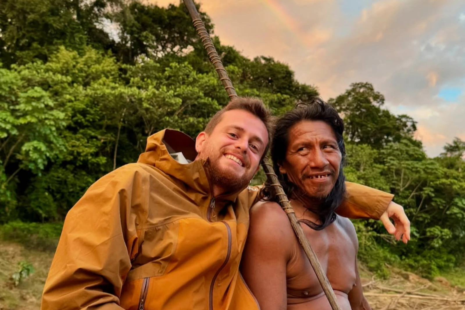 A Waorani elder with long hair, stretched earlobes, and a warm smile sits next to Alper Rende, who is wearing rain boots and making a peace sign.