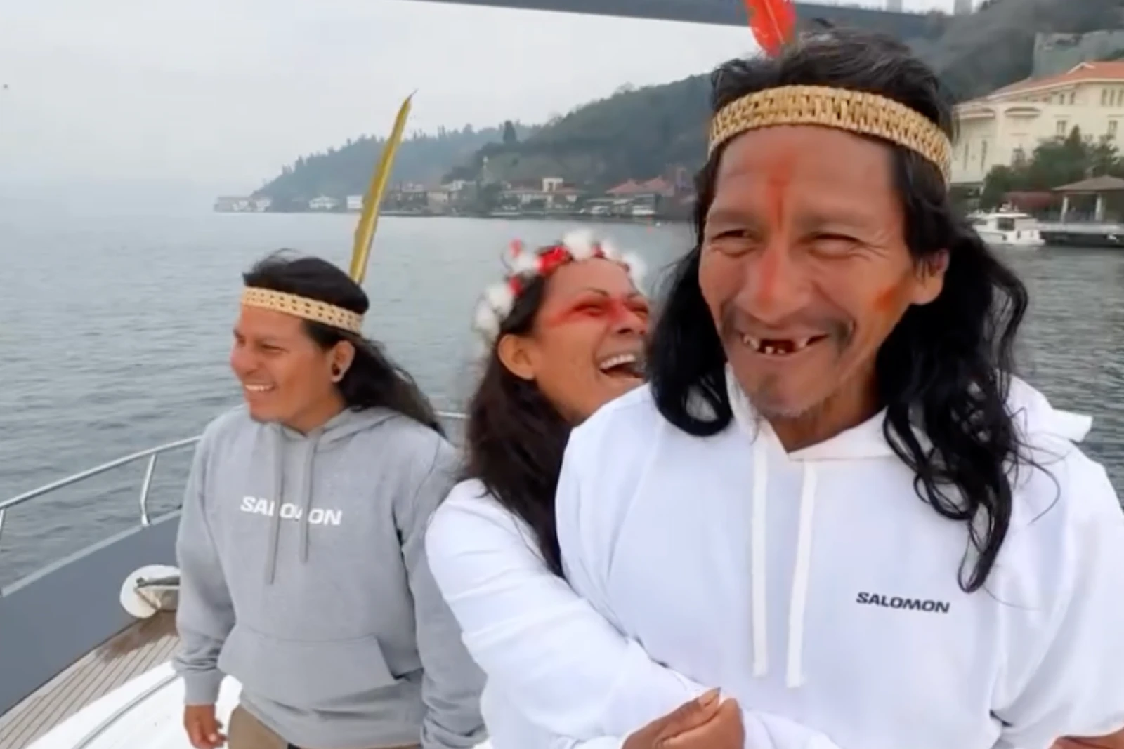 A Waorani woman and two men laugh together on a boat in Türkiye, wearing feathered headbands and white hoodies.