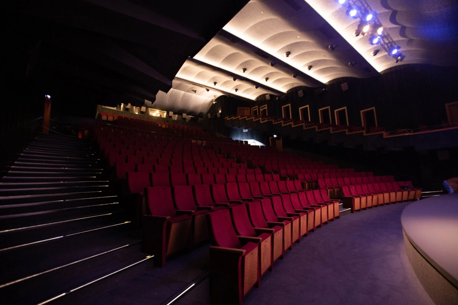 Interior view of the Atlas 1948 theater showing empty red velvet seats arranged in tiered rows. The auditorium features illuminated curved acoustic panels on the ceiling, lighted stairs along the sides, and a dark, intimate atmosphere