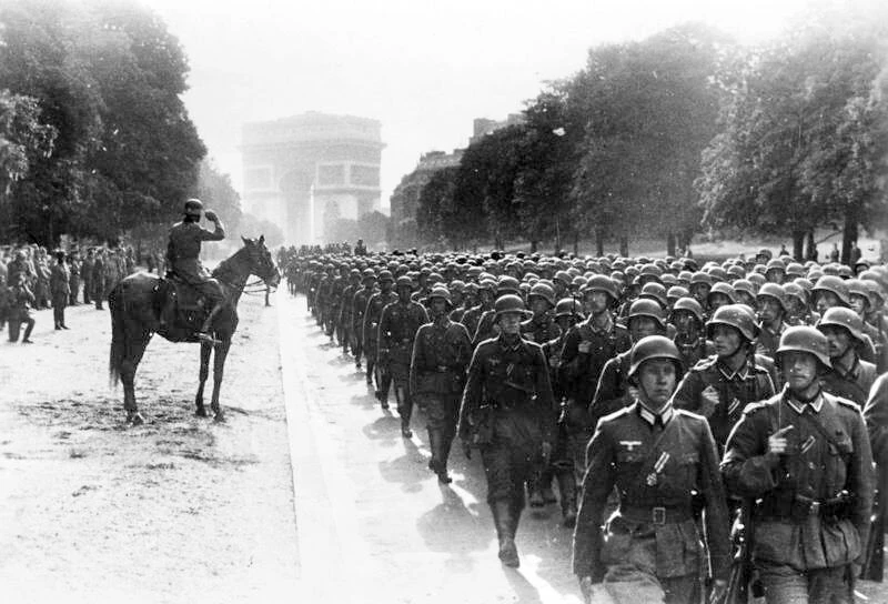 A black-and-white photograph of German soldiers marching in Paris during World War II, with a mounted officer saluting and the Arc de Triomphe in the distance.