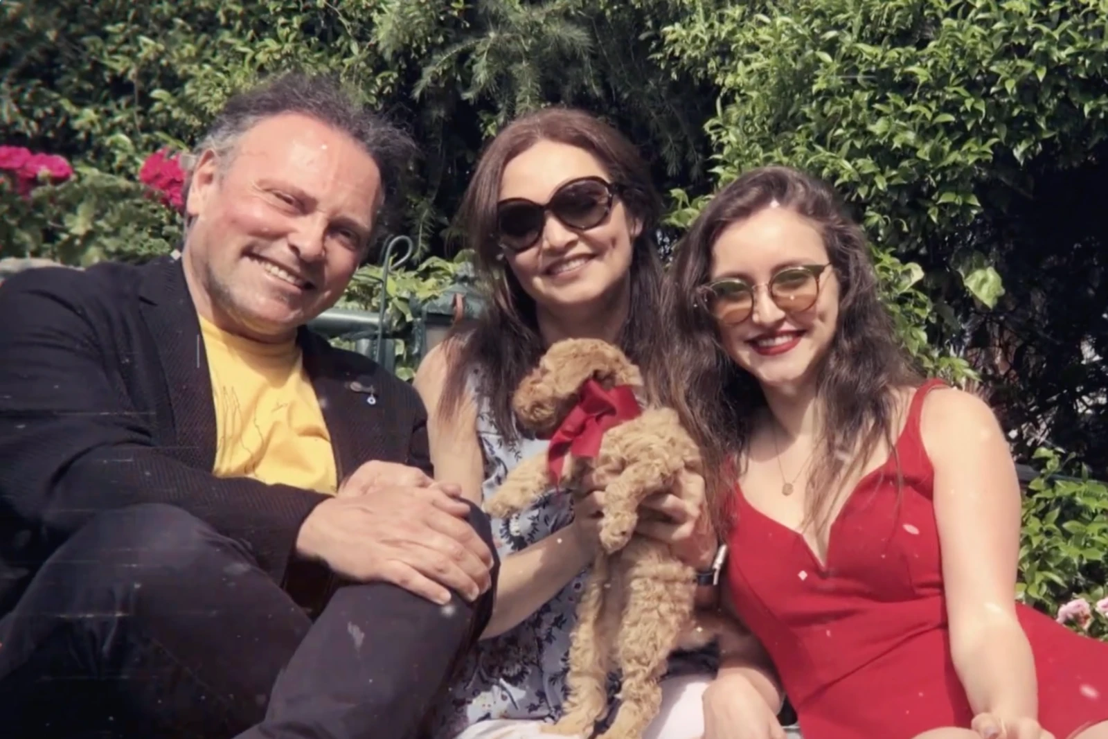 Ilayda Samilgil sits with her parents in a garden, holding a small dog with a red bow, all smiling at the camera.