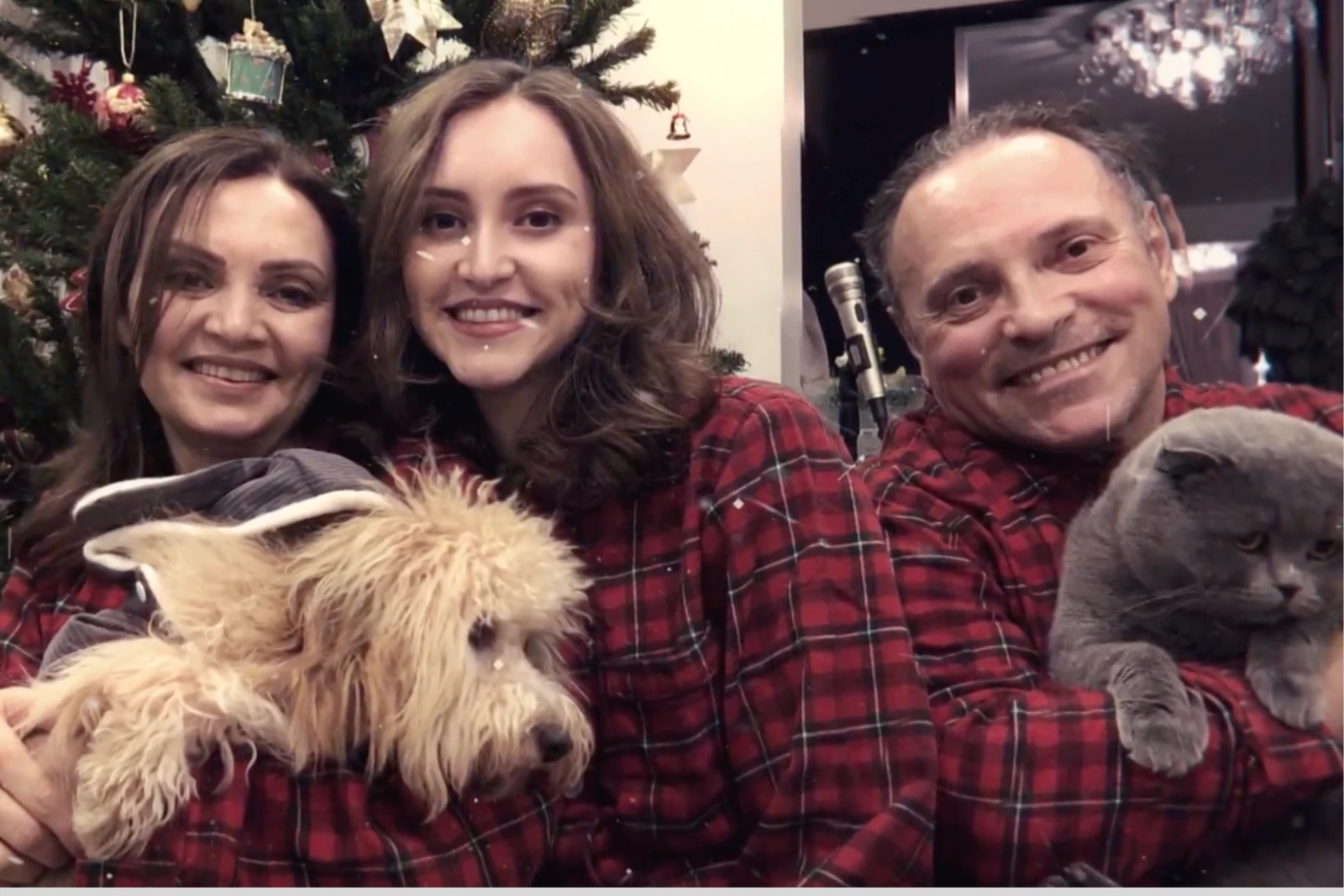 Ilayda Samilgil and her parents wear matching red plaid outfits, holding a dog and a cat in front of a Christmas tree.