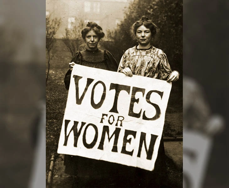 Two women from the early 20th-century suffrage movement stand holding a large ‘Votes for Women’ banner, advocating for women's right to vote.