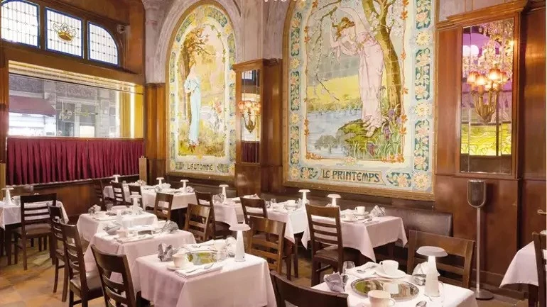 A well-lit dining area inside historical cafe Markiz Pastanesi, featuring neatly arranged tables with white tablecloths and elegant Art Nouveau murals depicting seasonal scenes on the walls.