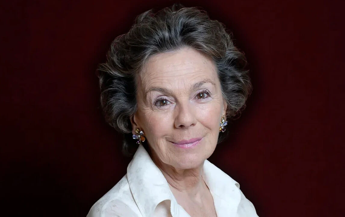 A portrait of Kenize Murad in a white blouse, wearing floral earrings, against a dark red background.