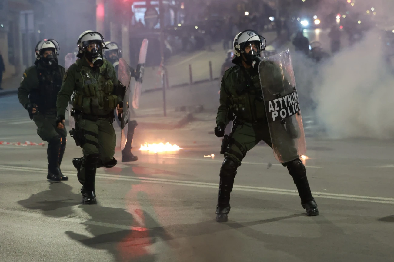 Riot police officers wearing gas masks and shields move forward amid smoke and flames during protests in Thessaloniki over the Tempi train accident.